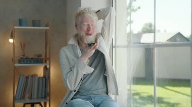 Olumlu genç albino adam cep telefonunu pencerenin yanındaki hoparlörden alıyor, evde pencere eşiğinde oturuyor. Albino adam gülümser, güler, rahatlar. Yüksek kalite 4k görüntü