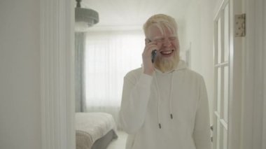 Geniş evde cep telefonuyla iletişim kuran albinizmli gülümseyen genç bir adam. Albino adam yatak odası, koridorlar, mutfağa geliyor. Beyaz iç mekan. Yüksek kalite 4k görüntü
