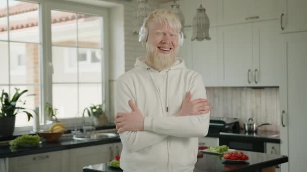 笑了笑 放松的白化病患者 戴着白色耳机站在厨房里 朝着节拍走去 阿尔比诺男人站在那里 双臂交叉 听音乐 高质量的4K镜头 — 图库视频影像