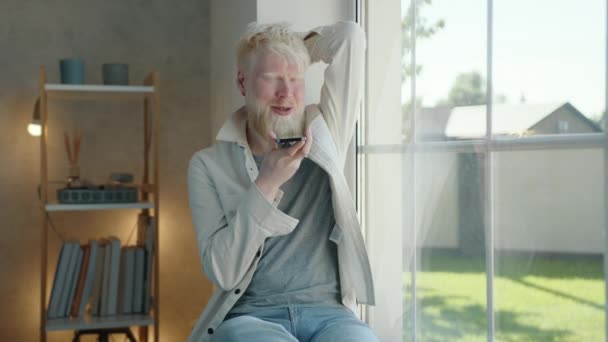 窓の近くのスピーカーフォンで携帯電話を手にしたポジティブな若いアルビノの男は 自宅の窓辺に座っている アルビーノの男は笑顔 リラックスしています 高品質4K映像 — ストック動画