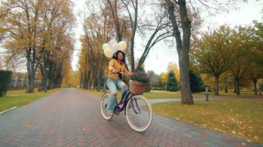 Hoş, siyah saçlı, kulaklıklı, parkta boş vaktini harcayan, elinde çiçekler ve balonlarla bisiklet süren, şarkı söyleyen, kıvırcık saçlı kadın. Yüksek kalite 4k görüntü