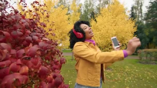中年时代的时髦 积极的卷曲女人 戴着红色耳机 享受着最喜爱的歌声 在秋天的公园里与红树 黄树和绿树共度时光 高质量的4K镜头 — 图库视频影像