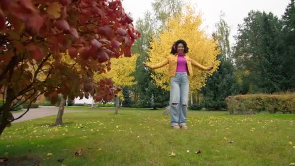 快乐的女性 有着短而黑的卷发 穿着撕破的牛仔裤 粉色的高领毛衣 黄色的夹克 戴着红色耳机歌唱着 秋天公园里的曲调飘扬着 高质量的4K镜头 — 图库视频影像