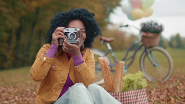 ショッピング サイクリング後の秋の公園の落ち葉の黄色い毛布に座ってビンテージカメラで写真を撮る魅力的な笑顔の色の女性 高品質の4K映像 — ストック動画