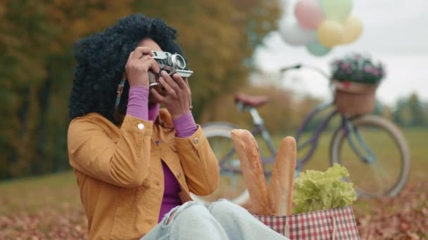 かわいい黒人女性が公園でレジャー時間を過ごし 落ち葉の黄色い毛布に座ってヴィンテージカメラで写真を撮ります ホビーさん サイクリング ショッピング 高品質の4K映像 — ストック動画