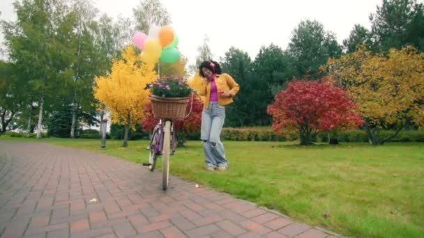 ヘッドフォン スタイリッシュな服チリン カラフルな風船と菊でバスケットで飾られた自転車の近くで踊る陽気なダークスキンレディ 高品質の4K映像 — ストック動画