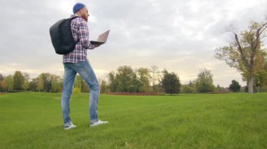 Genç erkek turist yeşil bir alanda geziniyor, sonbaharda çok renkli ağaçlar dizüstü bilgisayarında arama yapıyor. Gün batımında kasvetli bir sonbahar günü. Yüksek kalite 4k görüntü