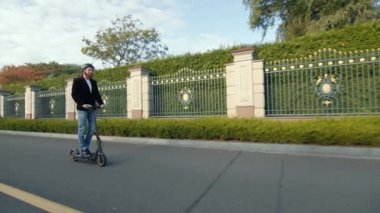 Parlak görünümlü, kot pantolon ve siyah ceketli bir sürücü elektrikli scooter ile uzun bir yol boyunca güzel metal ve ağaç çitlerinin yanından geçiyor. Sonbahar. Yüksek kalite 4k görüntü