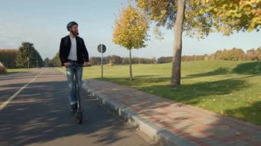 E-scooterlı neşeli adam yol boyunca seyahat ediyor, engin park alanının tadını çıkarıyor renkli sonbahar ağaçları, yeşil çimenler. Yüksek kalite 4k görüntü