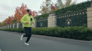 Spor giyimli genç bir erkek koşucu ve Bini asfalt yolda zarif metal çitlerin yanında çok renkli sonbahar ağaçları arkasında da bir sıra yeşil ağaçla koşuyor. Yüksek kalite 4k görüntü