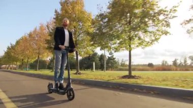 Sonbahar parkında güzel güneşli bir gün. Yeşil çimenler, çok renkli sonbahar ağaçları, sokak ışıkları, patikalar. Genç adam açık havada e-scooter sürmekten hoşlanıyor. Yüksek kalite 4k görüntü