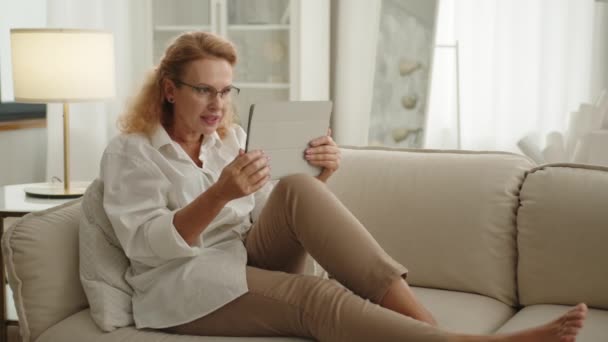 現代のライトルームの自宅に伸ばされた足で座っているかわいいブロンドの女性は 手でタブレットを握り ビデオチャットをしたり 感情的に話したり 手を振ったりします 高品質の4K映像 — ストック動画