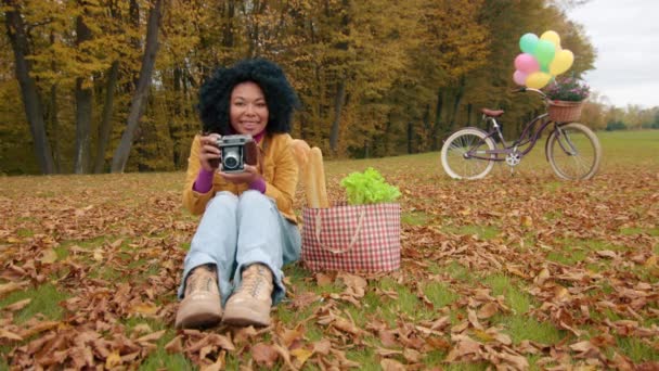 在秋天的公园里带着相机微笑可爱的女人 坐在草坪上给摄影师拍照 带着产品和装饰自行车的购物袋为背景 高质量的4K镜头 — 图库视频影像