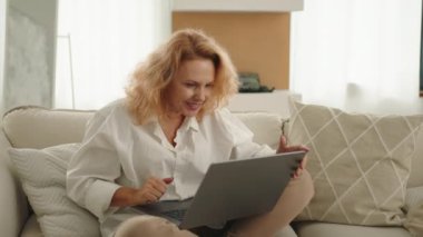 Beyaz tişörtlü, beyaz kollu ve bej pantolonlu mutlu orta yaşlı bir kadın merhaba diyor, görüntülü sohbet ediyor, dizlerinin üstünde dizüstü bilgisayar tutuyor. Yüksek kalite 4k görüntü