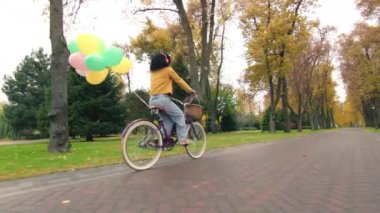 Yeşillik, sarı ağaçlar, çimenler, ıslak patikalar olan büyüleyici bir park alanı. Bisikletli bayanın arkadan görünüşü balonlarla, çiçeklerle süslenmiş. Yüksek kalite 4k görüntü