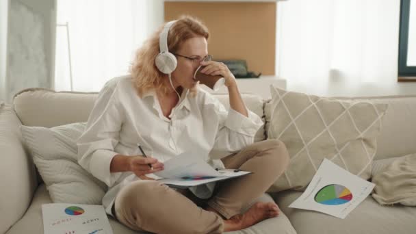ヨガに座っているかなり笑顔の女性は 家から仕事をし 音楽を聴き コーヒーを飲んで カラフルなパイチャートで紙をレビューする鉛筆を持っています 高品質の4K映像 — ストック動画