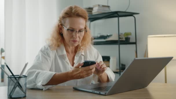 在现代公寓的一个舒适的房间里 一个戴着眼镜 戴着手提电脑的漂亮金发女人坐在餐桌旁 用手机输入信息 高质量的4K镜头 — 图库视频影像