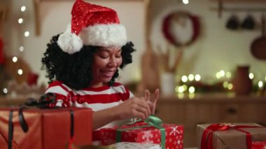 Gülümseyen orta yaşlı siyahi kadın rahat bir odada oturmuş Noel için hediyeler paketliyor, kutuyu kırmızı ambalaj kağıdıyla uzatıyor. Yüksek kalite 4k görüntü