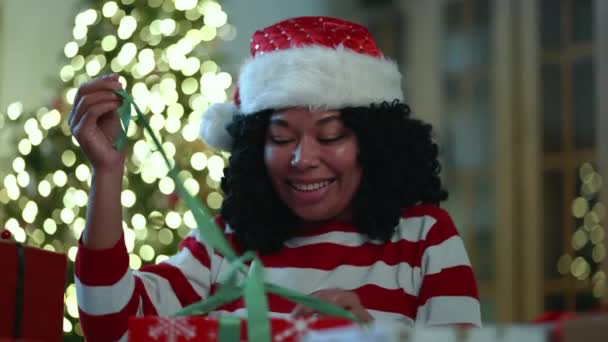 一个有着红白条纹长袖的快乐的黑人女人坐在一个房间里 房间里有大量礼物包裹 圣诞树模糊 有一个花环 高质量的4K镜头 — 图库视频影像