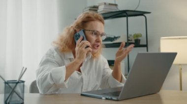 İş kadını masada oturuyor, cep telefonundan rahatsız edici bir şekilde dizüstü bilgisayara bakıyor. Sarışın kapanış aleti, sinir bozucu, bitirici bir konuşma. Yüksek kalite 4k görüntü