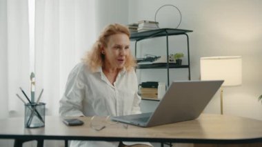 Kısa kıvırcık saçlı çekici bir kadın rahat bir odada oturuyor, dizüstü bilgisayarında yazıyor, heyecandan yumruklarını sıkıyor. Yüksek kalite 4k görüntü
