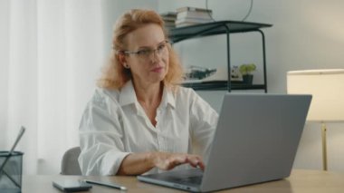 Güzel, bakımlı, gözlüklü sarışın kadın dizüstü bilgisayarla yazmayı bitiriyor, ışıkları yakıyor ve modern restorasyonlu bir odada iş yerini şekillendiriyor. Yüksek kalite 4k görüntü