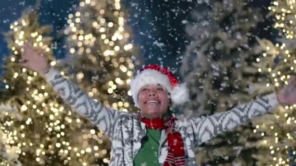 身穿针织羊毛衫的黑人妇女笑着庆祝冬天 向上看 在户外被雪花环绕 背景上点亮圣诞树 高质量的4K镜头 — 图库视频影像