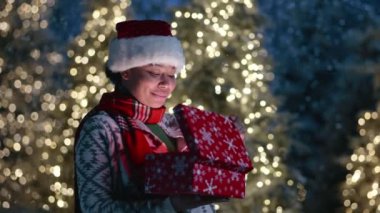 Boynunda kırmızı atkısı olan sevimli siyah kadın hediye paketine bakıyor, mutlulukla bağırıyor, aydınlık Noel ağaçlarıyla dışarıda duruyor, kar yağıyor. Yüksek kalite 4k görüntü