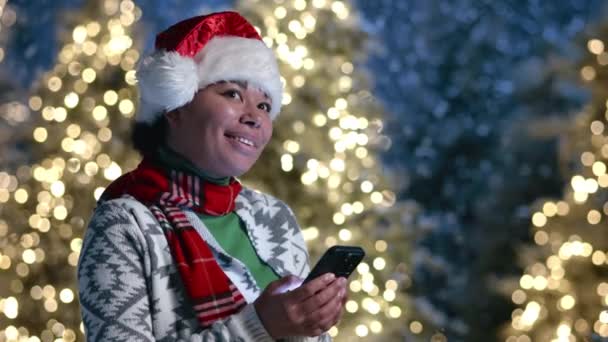 穿着羊毛衫和红围巾的非洲美女看着手机 笑着站在室外 模糊的背景照明的圣诞树和陷阱 高质量的4K镜头 — 图库视频影像