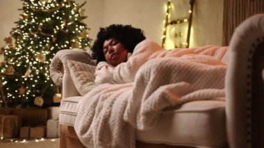 Beyaz pijamalı kıvırcık siyah kadın, sarı çelenkli Noel ağacıyla süslenmiş bir oturma odasında, ekoseyle kaplı kanepede uyuyor. Yüksek kalite 4k görüntü