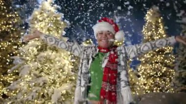 Örgü hırka giyen mutlu esmer kadın kar yağışı, fırıl fırıl dönen, el kaldıran, zıplayan. Arka planda aydınlatılmış xmas ağaçları. Noel havası. Yüksek kalite 4k görüntü
