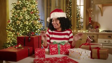 Noel Baba şapkalı gülümseyen siyah kadın, çizgili uzun kollu neşeli bir mutfakta çelenklerle süslenmiş, ışıklandırılmış xmas ağacı, hediye paketlerine bayram eden. Yüksek kalite 4k görüntü