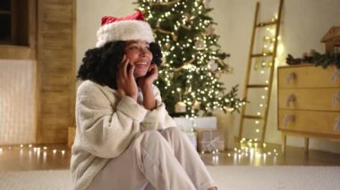 Beyaz pijama ve Noel şapkası içinde, parlak kostüm ağaçları, hediyeler ve çelenklerle dolu rahat bir odada yerde oturmuş gülümseyen kıvırcık, genç, siyah bir kadın. Yüksek kalite 4k görüntü