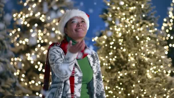サンタの帽子の陽気なアフリカの女性は 豪華な雪で装飾された公園に座って 息子が贈り物を持って箱を与えている間 照らされたクリスマスツリー 高品質の4K映像 — ストック動画