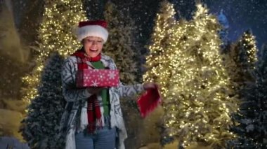 Noel Baba şapkası takmış, hırka örmüş, karlı bir gece parkında elinde parlak Noel ağaçlarıyla hediye paketi tutan güzel siyahi bir kadın. Xmas hediyesi. Yüksek kalite 4k görüntü