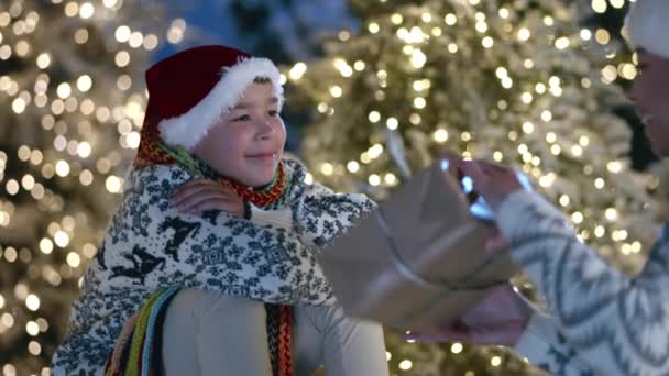 10岁的儿子坐在一个美丽的地方 有模糊的圣诞树和照明 当非洲女人 妈妈带着礼物来 拥抱男孩 高质量的4K镜头 — 图库视频影像
