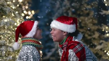 Sıcak süveter ve eşarp giymiş, dışarıda el çırpmaca oynayan, neşeyle burunlarına dokunan çocuklu mutlu bir anne. Çiçekli bulanık Noel ağaçları. Yüksek kalite 4k görüntü