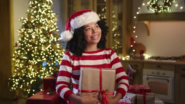 サンタの帽子にかわいい黒人女性を興奮させ 赤いリボンで金の紙に包まれた大きな箱を抱擁する白い赤い縞の長袖 イルミネーションされたXmasツリー 高品質の4K映像 — ストック動画