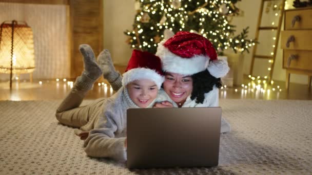 快乐的黑人女人 戴着圣诞帽的小男孩躺在地板上 笑着看着笔记本电脑 舒适轻盈的现代客厅 有照明 圣诞树 高质量的4K镜头 — 图库视频影像