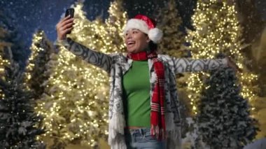 Hırkalı, kot pantolonlu, kırmızı eşarplı neşeli bir Afrikalı kadın merhaba diyor, gülüyor, yoğun kar yağarken cep telefonundan video görüşmesi yapıyor, çelenkli kostüm ağaçları var. Yüksek kalite 4k görüntü