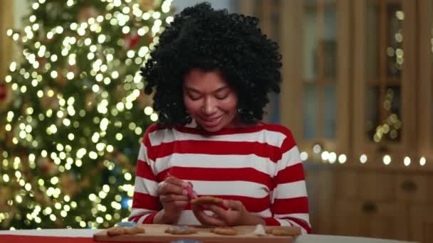 中年的非洲裔美国女士装饰着圣诞饼干 观望着烘焙着烘焙着的面包店 坐在餐桌旁 背后点缀着明亮的圣诞树 高质量的4K镜头 — 图库视频影像