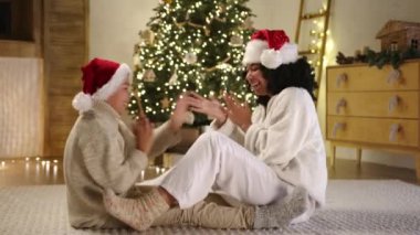 Noel arifesi. Noel Baba şapkalı mutlu anne ve oğlu ve sıcak ev kıyafetleri içinde parlak kostüm ağacıyla aydınlık bir odada, dekoratif merdivende çelenk ile oynuyorlar. Yüksek kalite 4k görüntü