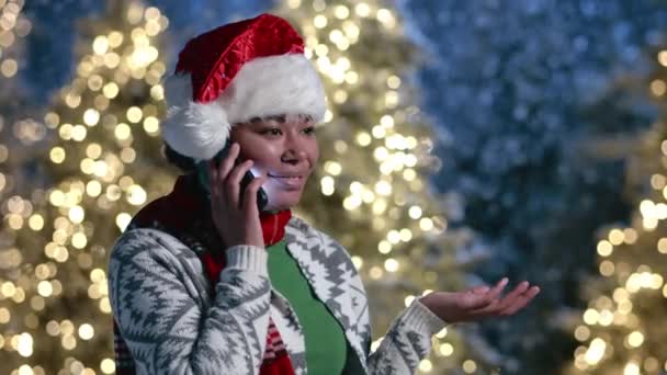 穿着开襟羊毛衫 戴着装饰品 戴着红色围巾的年轻黑人女子 高兴极了 她站在室外用手机说话 有照明的圣诞树 高质量的4K镜头 — 图库视频影像