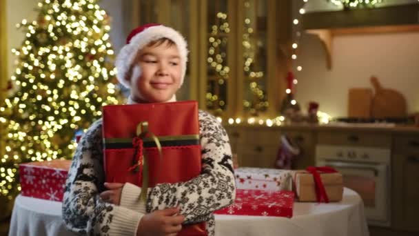 リボンが付いている赤い包むペーパーの大きい箱を握る暖かい装飾されたセーターの夢のような9 10歳の子供 輝くガーランドの美しいクリスマスツリー 高品質の4K映像 — ストック動画