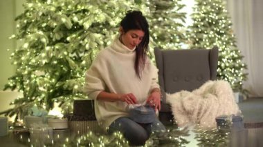 Kafkasyalı, rahat kıyafetli bir kadın Noel süslemeli bir evde hediye kutusunu sarar. Genç bayan yeni yıl arifesinde hediyeler hazırlar. Yüksek kalite 4k görüntü