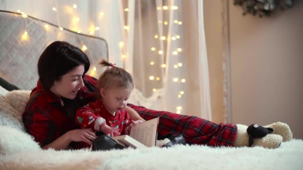 母亲和女儿穿着圣诞服装看书 白人妇女和她的孩子躺在床上 墙上挂着圣诞彩灯 高质量的4K镜头 — 图库视频影像