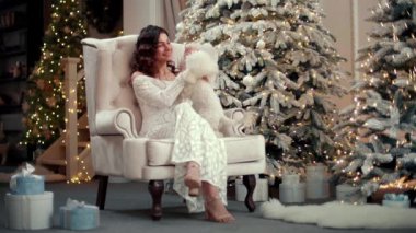 Beyaz elbiseli bir kadın Noel ağacında evcil hayvanıyla oynuyor. Şenlikli bir iç mekanda sosyetik bir kız bir köpekle oynuyor. Yüksek kalite 4k görüntü