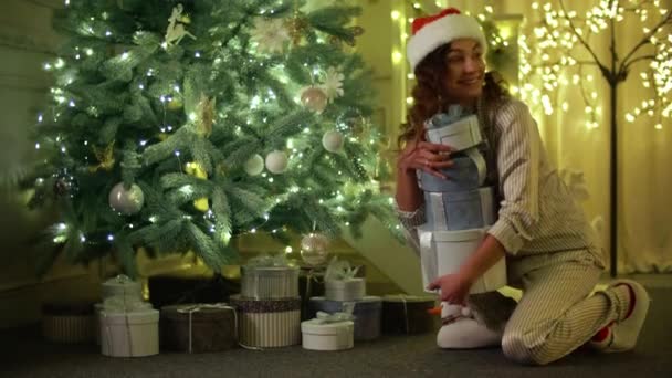 サンタクロースの帽子をかぶった陽気な女性がクリスマスツリーの下にプレゼントを隠します パジャマを着た若い女性がクリスマスの準備をします 高品質4K映像 — ストック動画