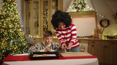 Çelenkler, Noel çelenkleri ve ışıklandırılmış Noel ağacı ile süslenmiş modern ışık mutfağı. 9-10 yıllık anne ve oğlu zencefilli kurabiyelere bakıyor. Yüksek kalite 4k görüntü