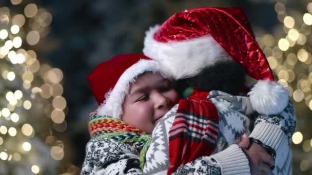 暖かい装飾されたセーターで9 10年の幸せな少年 サンタの帽子と赤いチェッカーされたスカーフで愛する母親と結ばれたスカーフ 輝くイルミネーション 高品質の4K映像 — ストック動画